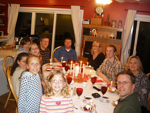 A multi-generational family gathered around an oval shaped dining table that is set with formal place settings, candles, and wine glasses.